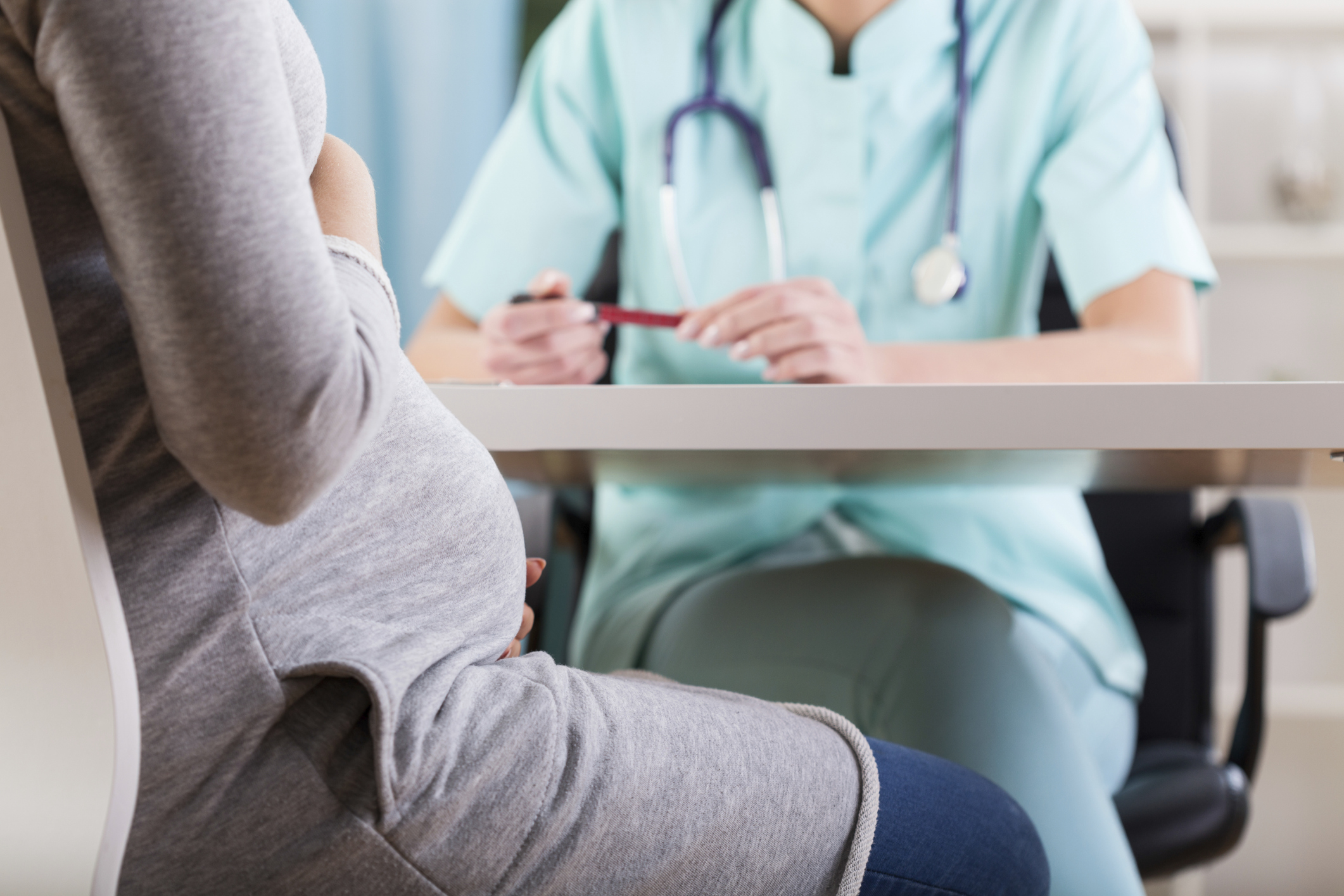 Mulher grávida vestida de cinza e mangas compridas em consulta com médica. Não é possível ver o rosto delas, estão sentadas.