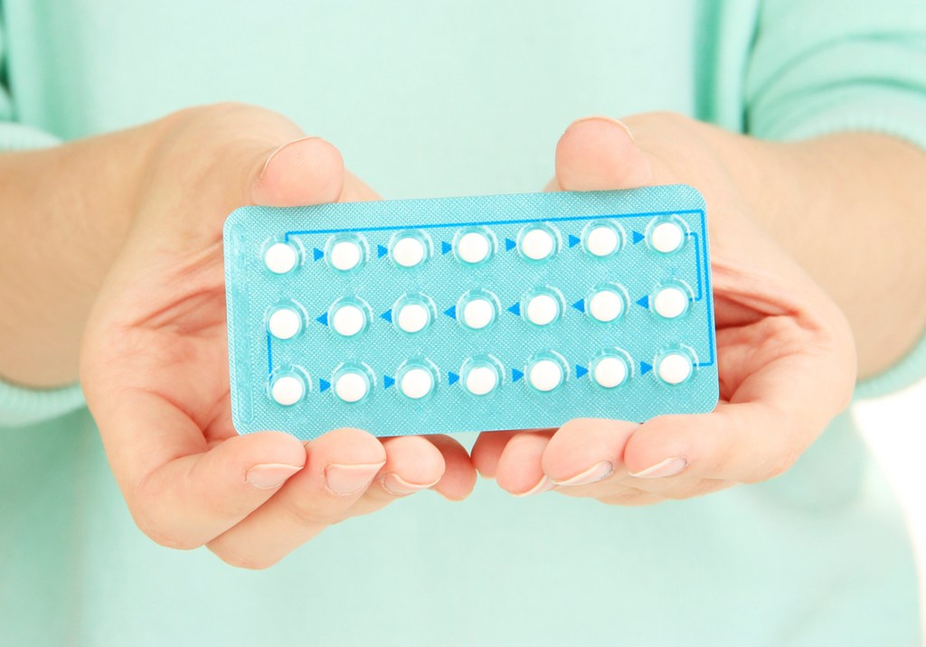 Mãos de pele clara femininas segurando uma cartela azul de pílulas brancas.
