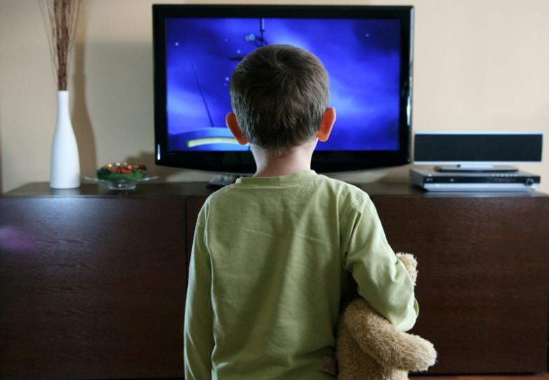 A boy holding a teddy bear looks at the television screen