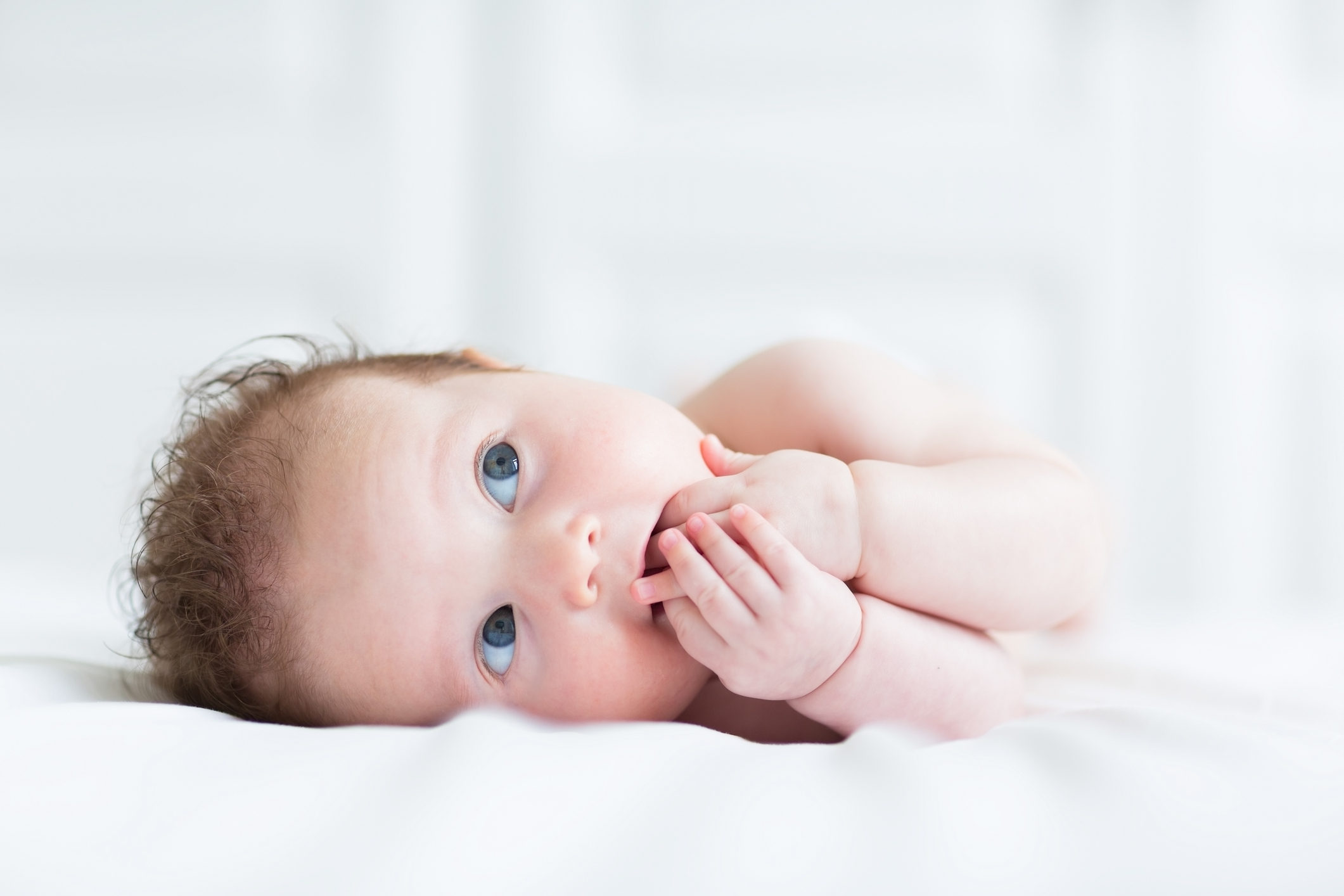 bebê de pele branca, cabelo castanho claro e olhos azuis deitado de lado, em cama com lençol branco. Ele está com as duas mãos na boca e olha para cima.