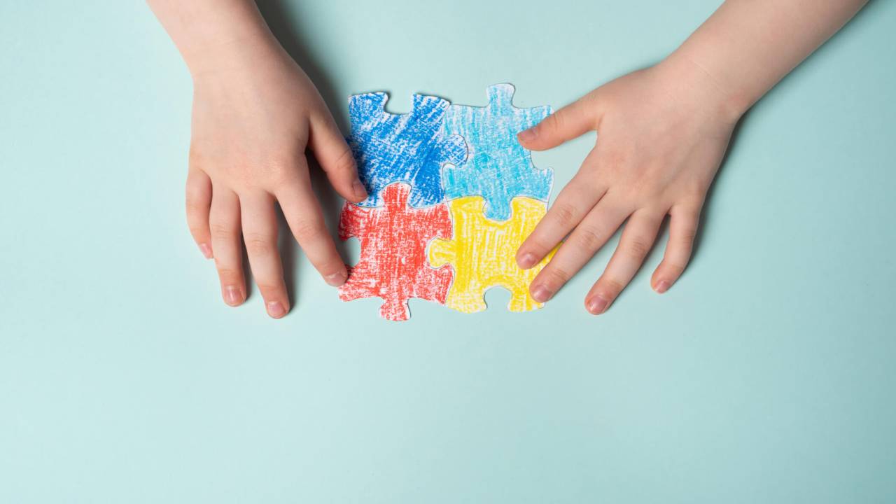 Sobre uma superfície azul clara, duas mãos infantis de pele clara montam quatro peças de um quebra-cabeça feito de papel e pintado a mão. Cada peça de uma cor: azul escuro, azul claro, vermelho e amarelo. Elas já estão unidas.