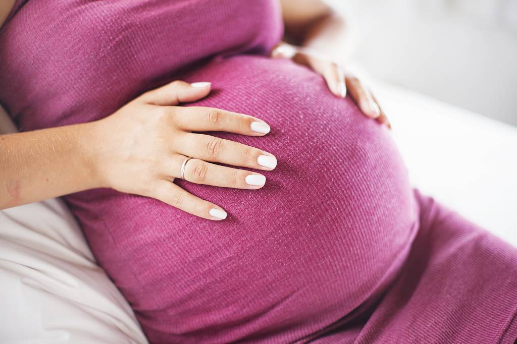 Grávida sentada no sofá com a mão na barriga. Ela usa um vestido rosa escuro e tem a pele clara. Não é possível ver o rosto nem as pernas.