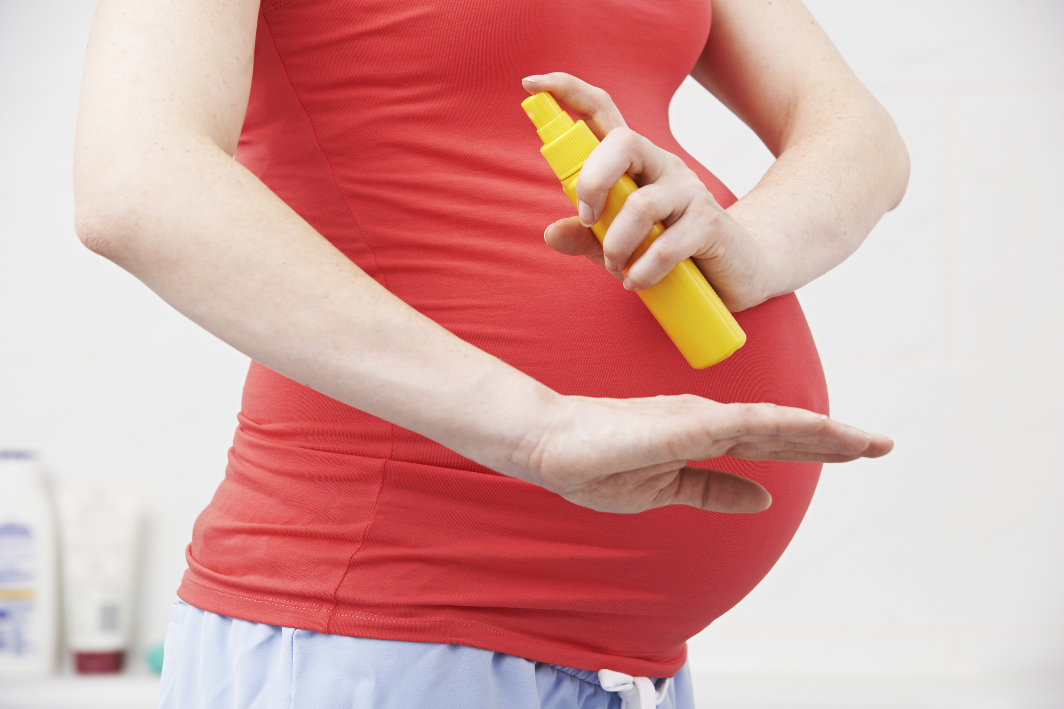 Mulher grávida aplicando repelente. Na imagem, aparece do peito até a cintura da mulher de pele branca. Ela veste uma camiseta vermelha, segura um frasco amarelo e espirra o produto em um dos braços.