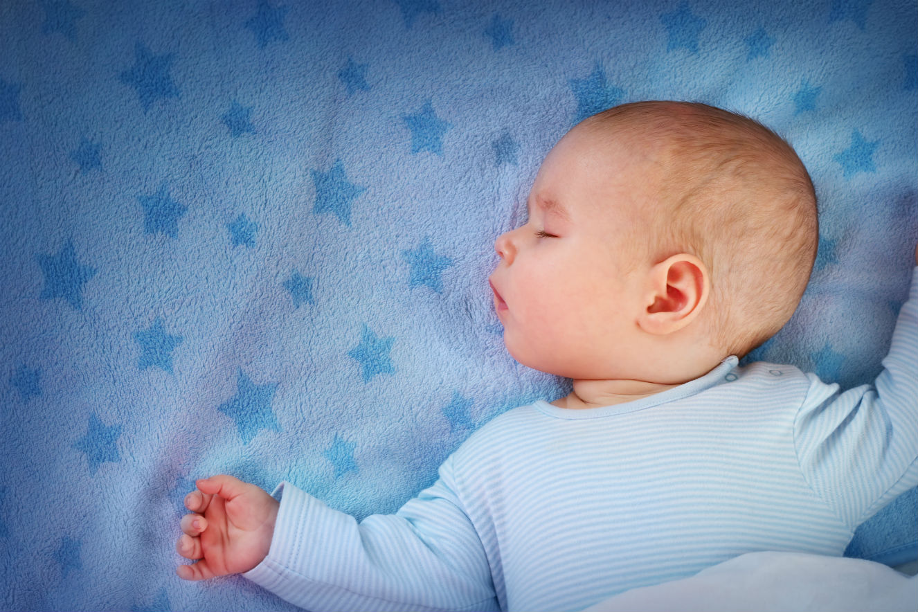 Bebê dormindo sobre lençol azul de estrelas azuis. O bebê tem pele clara, usa uma roupa azul clara, está de olhos fechados e com o rosto virado de lado.