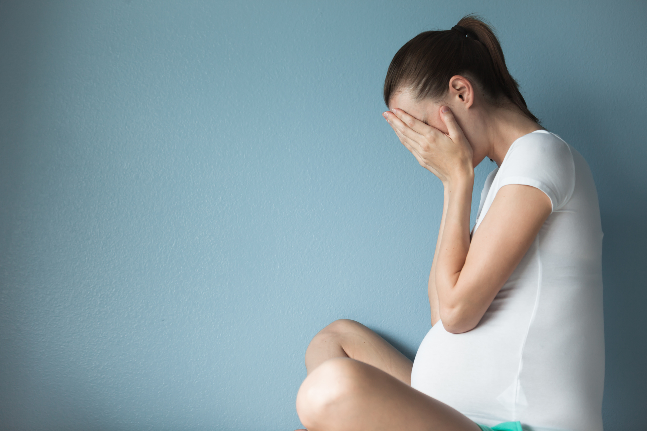 Mulher grávida sentada no chão de perfil, com o rosto escondido pelas mãos, como se estivesse chorando. Ela veste uma roupa toda branca. O fundo é azul.