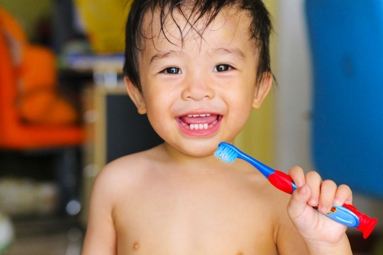 Menino asiático, por volta de 2 anos, escovando os dentes com uma escova azul. Ele está sorrindo e sem camisa.