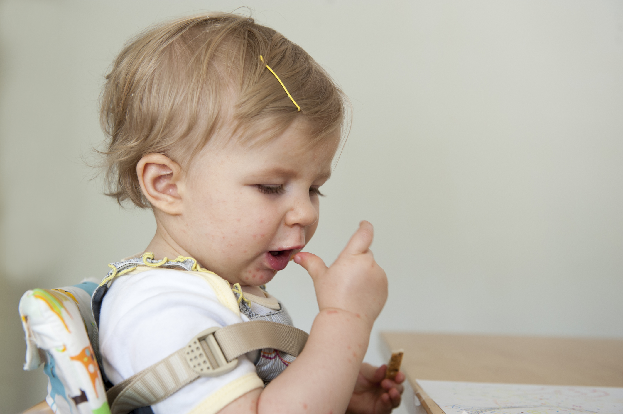 criança com por volta de 1 ano, menino, pele clara e cabelo loiro, sentado na cadeirinha de alimentação. Está levando um dedo a boca.