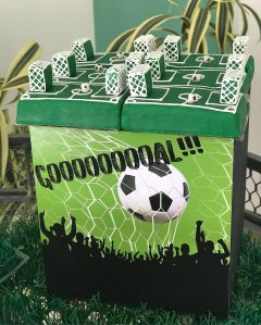 Bolo em formato de caixa, em que o topo lembra um jogo de tabuleiro de futebol, inspirado em Copa do Mundo