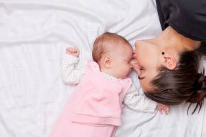 Mãe dando um beijinho na testa da filha que está dormindo de barriga para cima