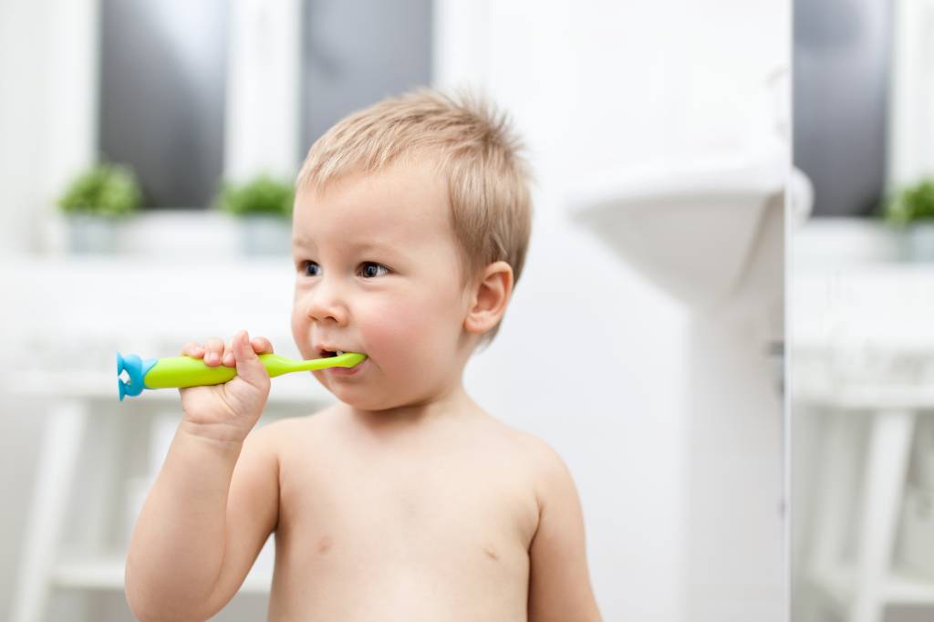 Bebê menino, por volta de 2 anos, escovando os dentes. É branco, loiro e com os olhos castanhos. Está segurando sozinho uma escova de dentes verde, que está na boca. Está sem camiseta e aparece da barriga para cima. O fundo da foto é desfocado e branco.