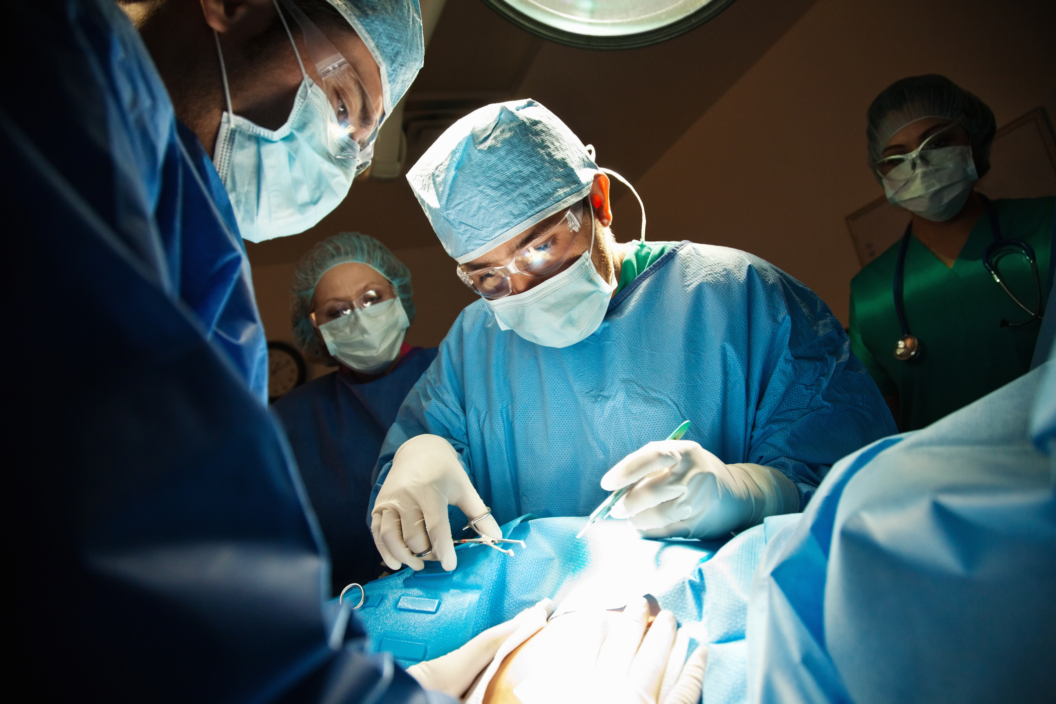 cirurgia cesariana. Na foto, é possível ver um médico manipulando instrumentos na mesa de cirurgia e outros profissionais ajudando. Estão usando roupa de proteção azul, máscara e touca.