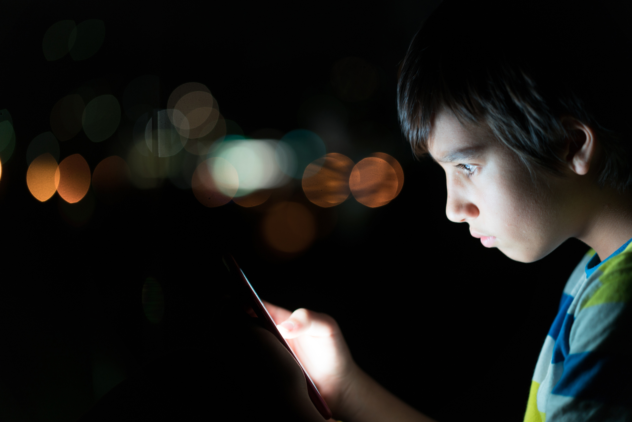 Criança usando o celular no escuro. O rosto do menino, que tem por volta de 8 anos, está iluminado pela tela.