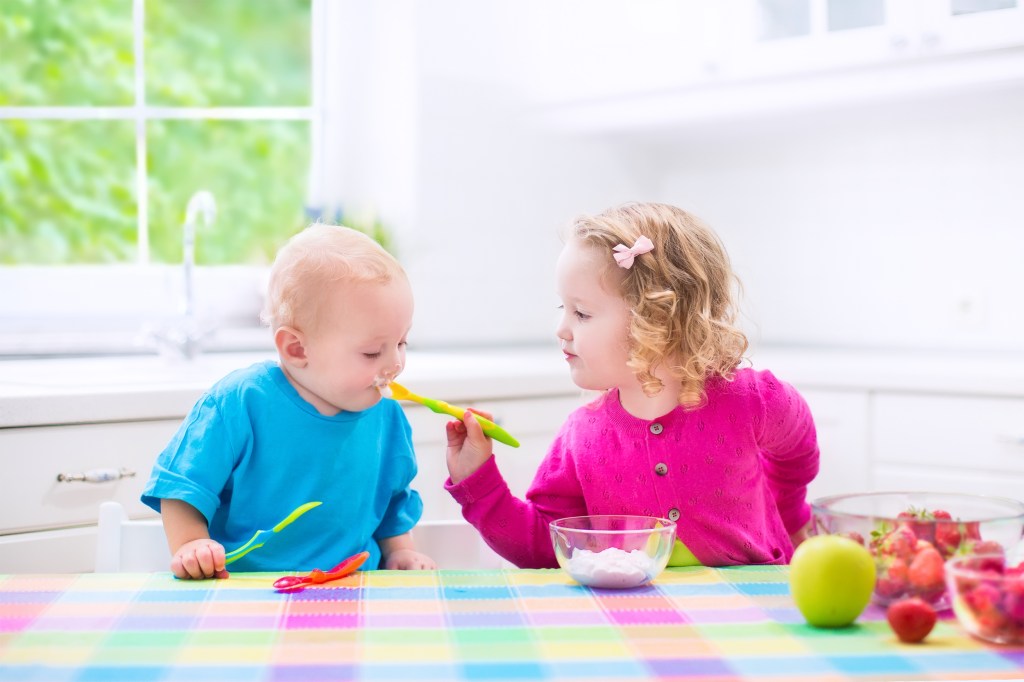 Na foto, há duas crianças brancas e loiras. Um menino, à esquerda, com menos de 1 ano, de camiseta azul. Uma menina, à direita, com por volta de 4 anos, de camiseta pink. Eles estão à mesa e a menina dá uma colherada de iogurte para ele.