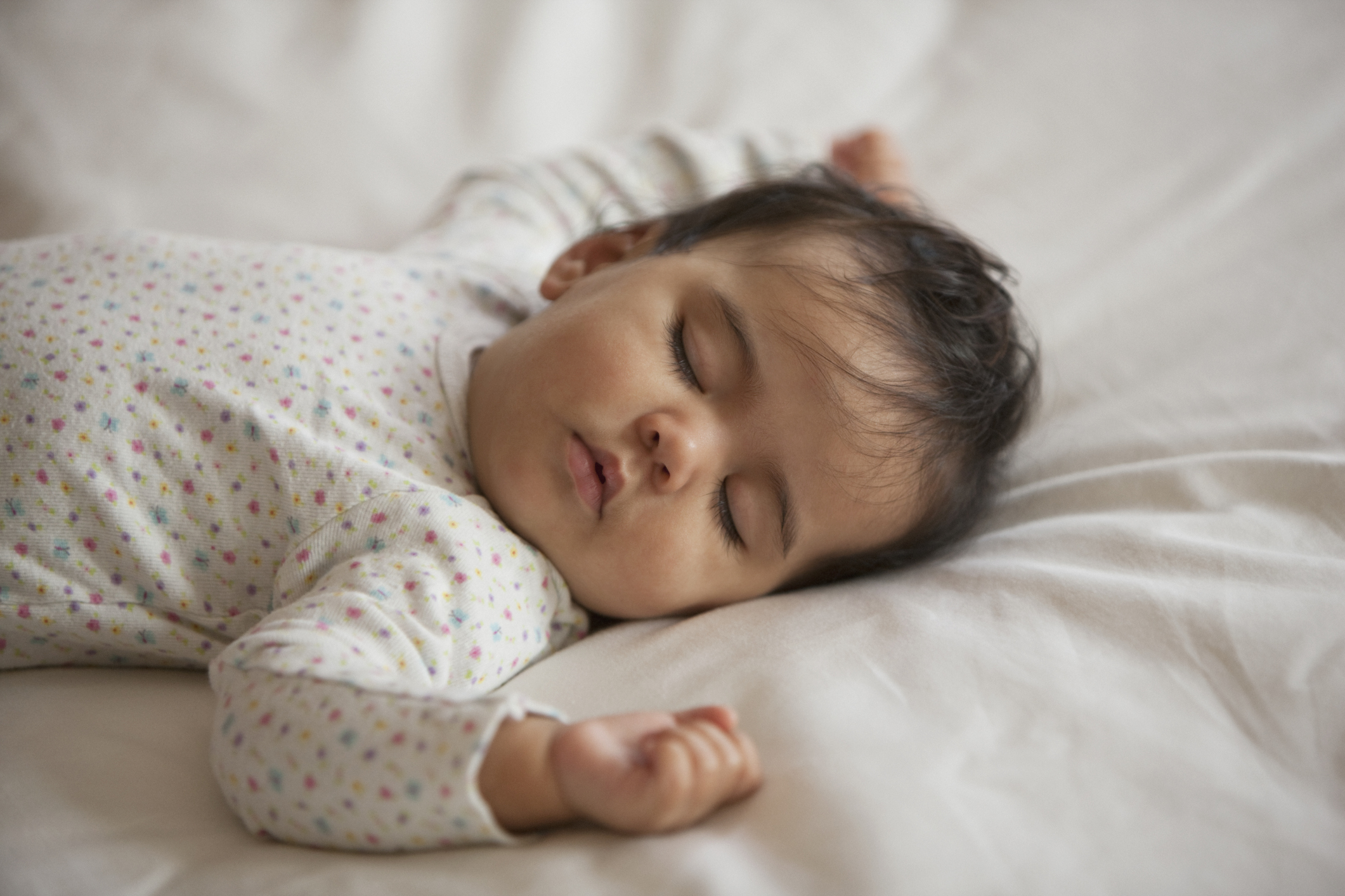 Bebê vestido de branco, pele clara, cabelo castanho escuro, dormindo de barriga para cima com os bracinhos abertos sobre colchão com lençol branco.