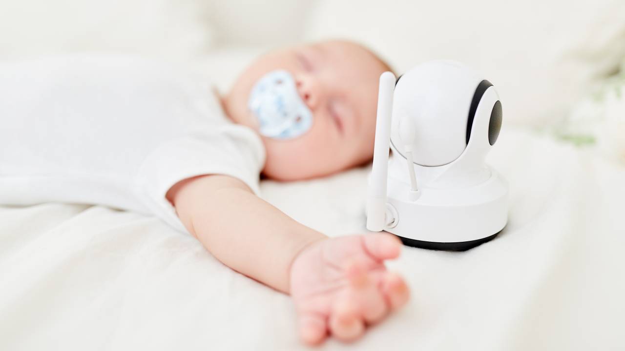 Babá eletrônica. A câmera está sobre o colchão, ao lado de um bebê dormindo, chupando chupeta, de barriga para cima e com os braços abertos.