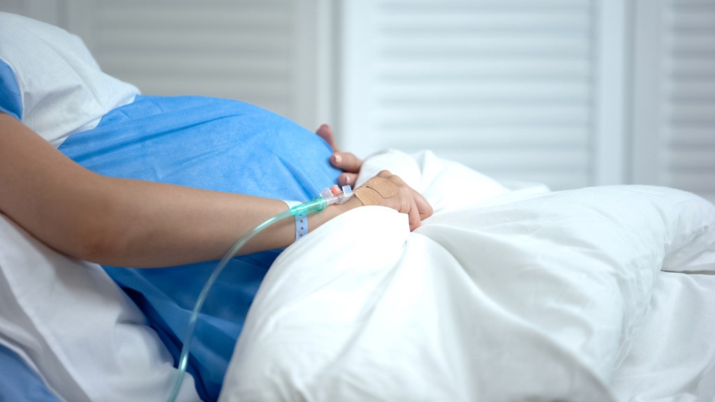 Na foto, é possível ver parte do corpo de uma mulher grávida deitada em cama de hospital. Ela usa roupa hospitalar azul, está com as mãos em baixo da barriga grande, com um tubo conectado à mão direita. Ela está coberta por um lençol branco.