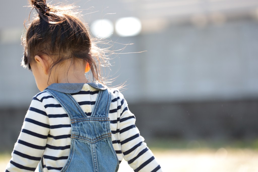 criança pequena de costas, com o cabelo castanho preso, usando camisa listrada e jardineira jeans