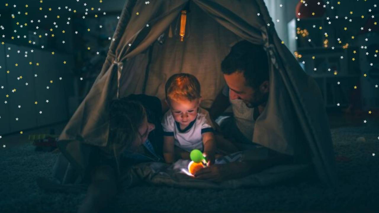 Mãe, pai e bebê no meio dos dois, dentro de uma cabaninha, com a luz principal apagada e uma lanterna acesa. Eles estão lendo um livro.