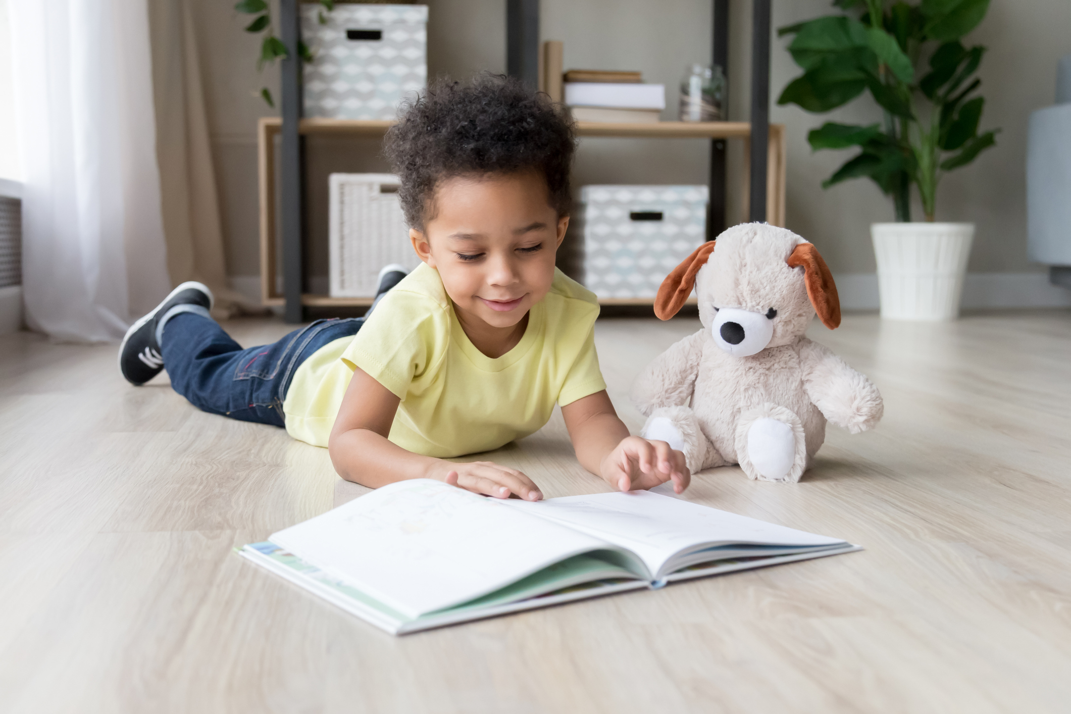 Criança pequena lendo livro sozinha deitada de barriga no chão. É um menino usando calça jeans e camiseta amarela, tem os cabelos pretos, curtos e crespos. Ao lado dele, há um cachorro de pelúcia sentado.