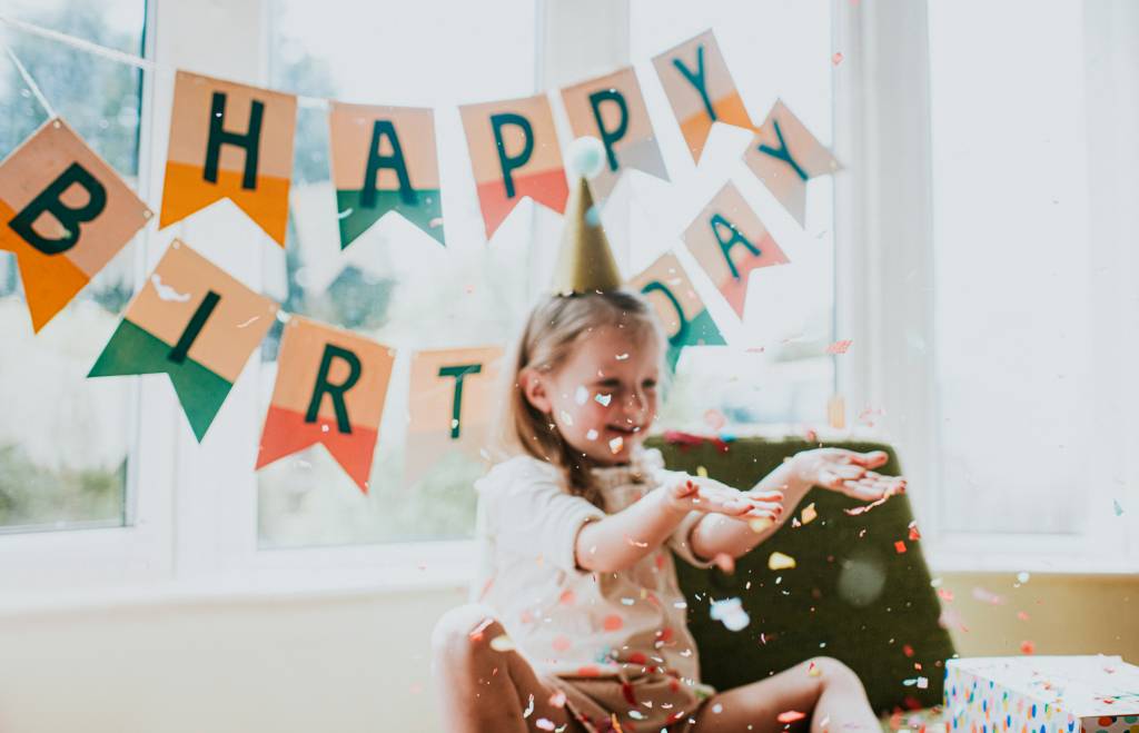 criança com chapéu de aniversário jogando confetes para o alto. Atrás dela, bandeirinhas formam a expressão Happy Birthday, Feliz Aniversário em inglês