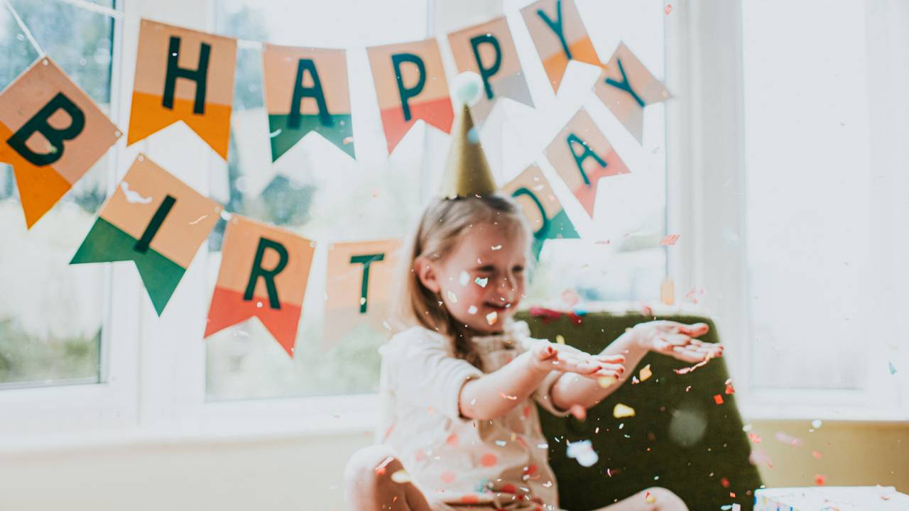criança com chapéu de aniversário jogando confetes para o alto. Atrás dela, bandeirinhas formam a expressão Happy Birthday, Feliz Aniversário em inglês