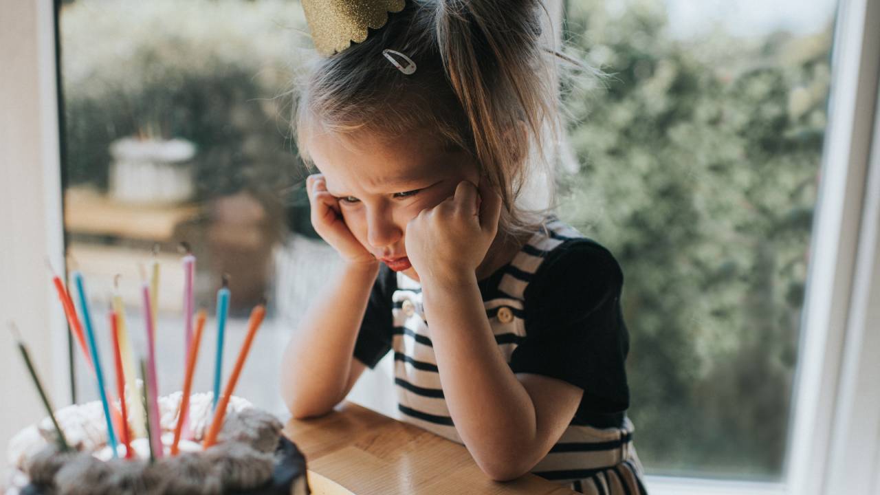 Menina triste diante do bolo de aniversário. A garota tem o cabelo preso, usa um chapéu de papelão na cabela e veste uma camiseta preta de mangas curtas com um vestido listrado preto e branco. Há velas coloridas sobre o bolo.