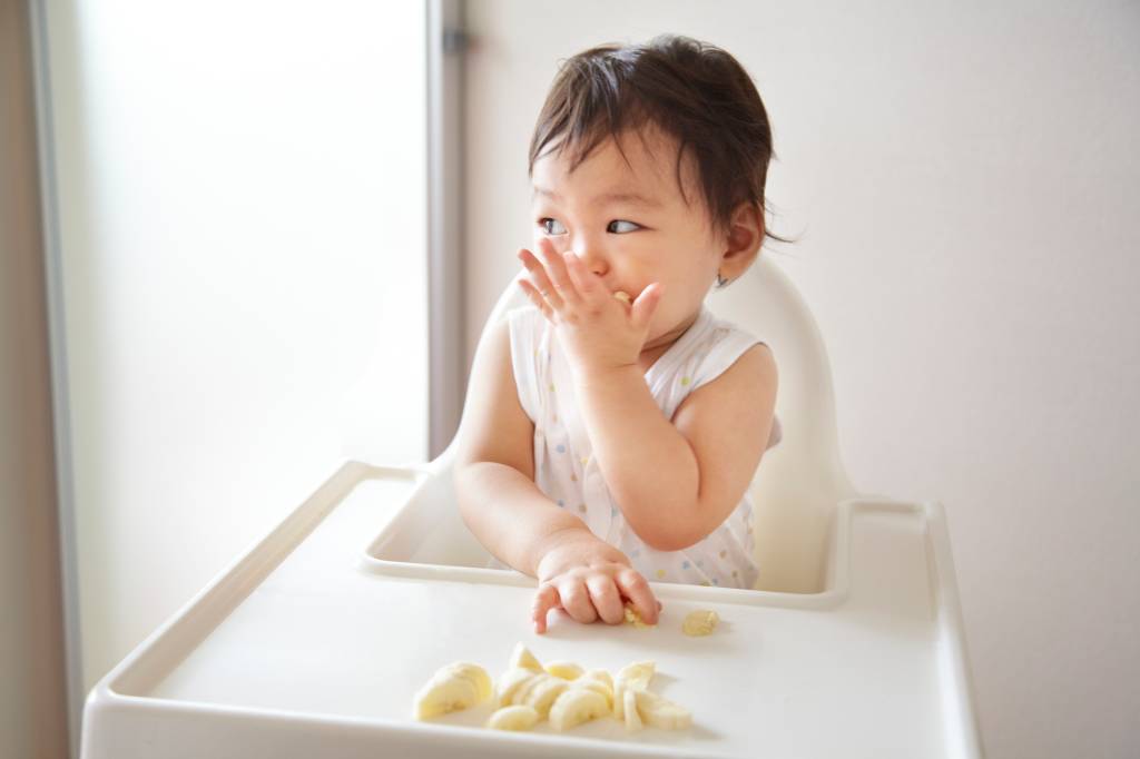 bebe-introdução-alimentar criança sentada em uma cadeira de alimentação comendo uma fruta que está sobre a mesa acoplada no acessório