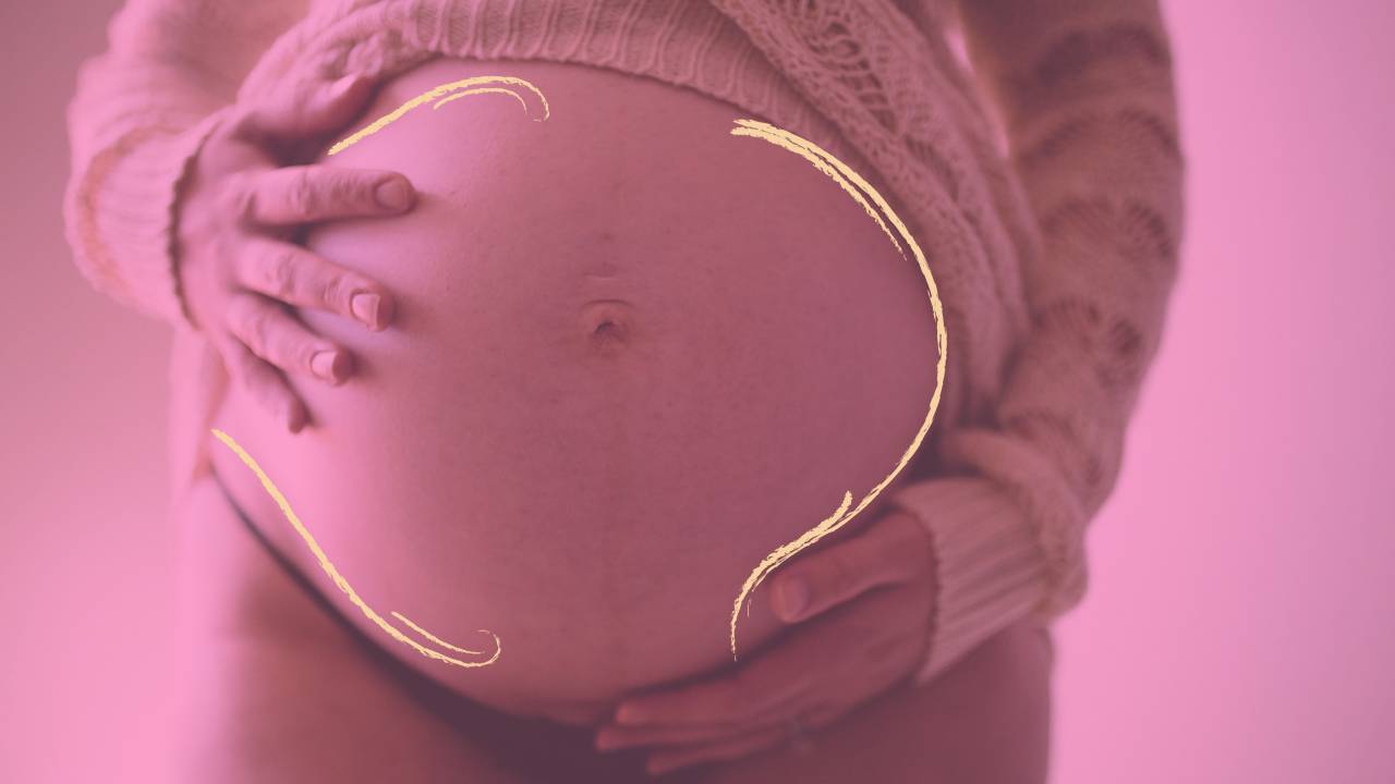 Na foto, há uma grande barriga de grávida, com as duas mãos sobre ela. A blusa, de lã, está levantada, deixando a barriga de fora. Há um filtro rosa sobre a imagem.