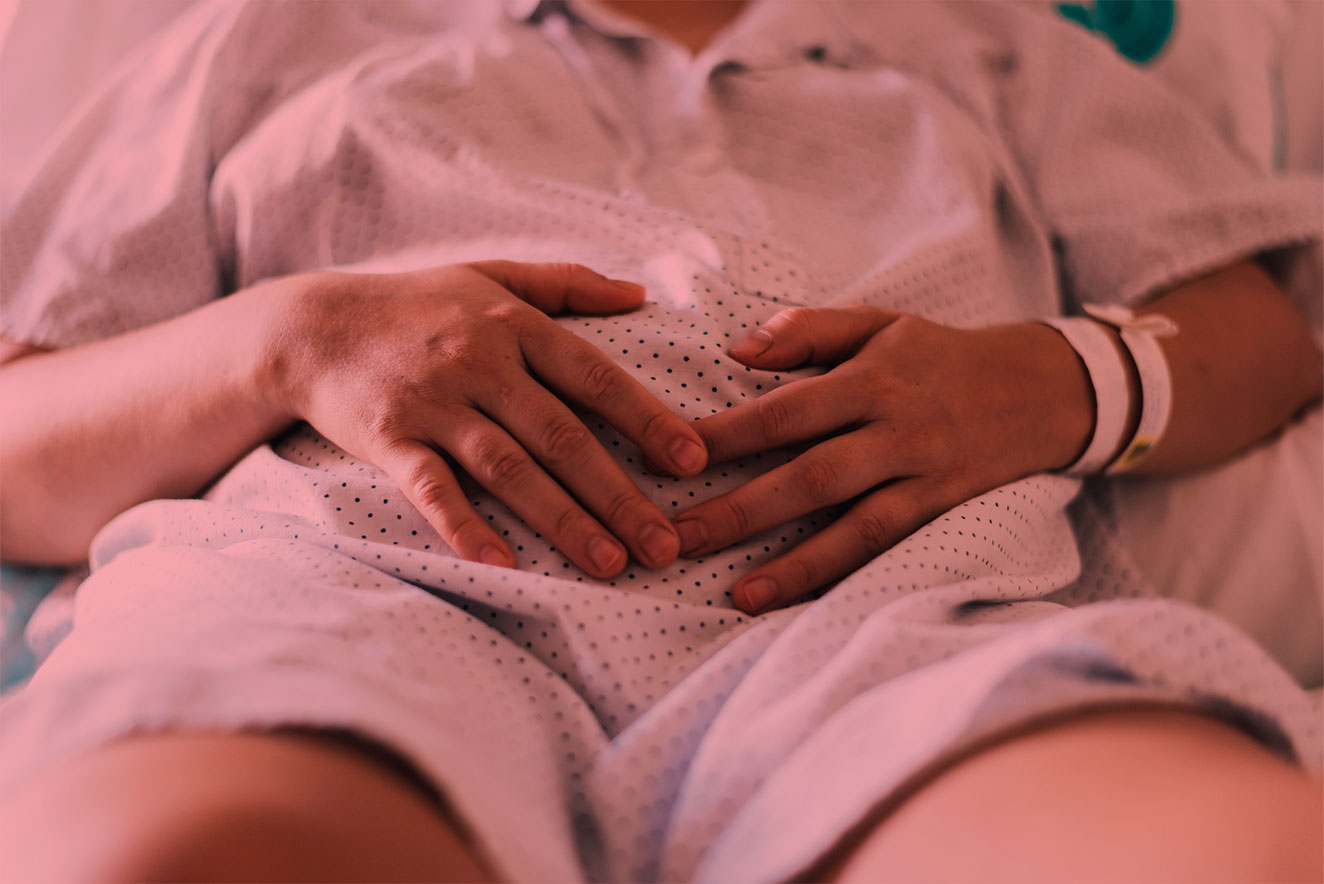 Mulher em hospital, usando roupa hospitalar, deitada e com as duas mãos sobre a barriga. Há um filtro vermelho sobre a foto.