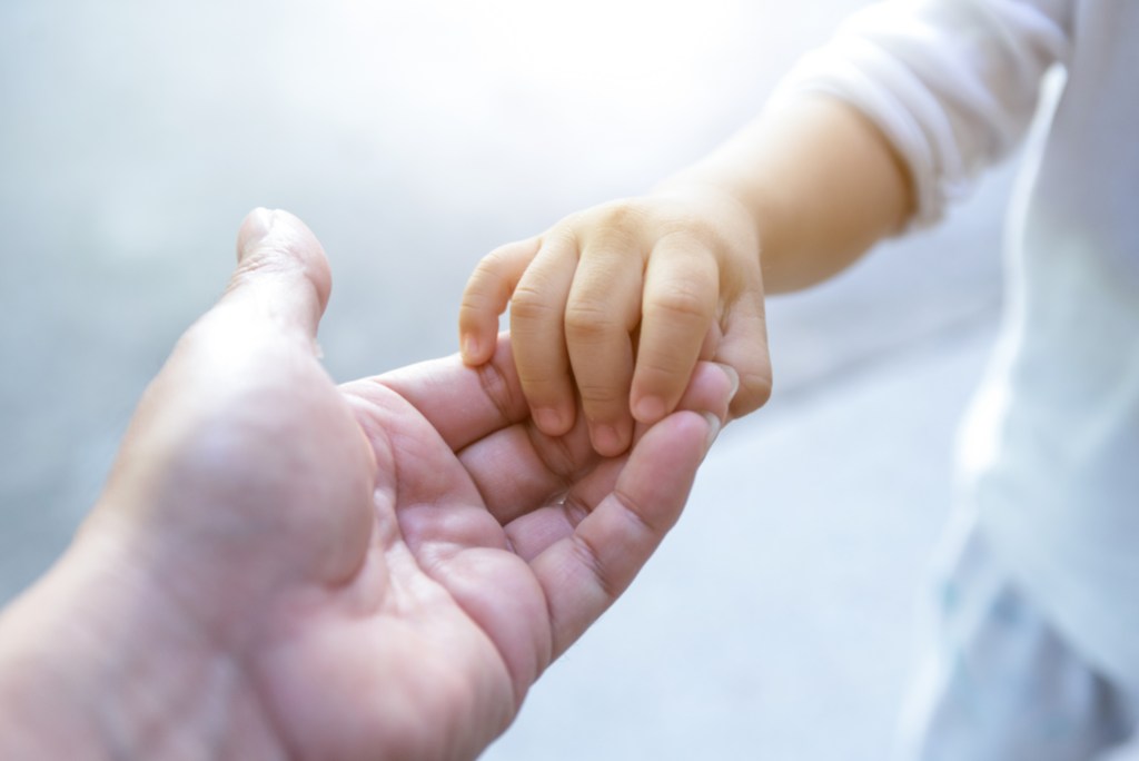 Na foto, uma mão infantil segurando uma mão adulta, ambas de pele branca.