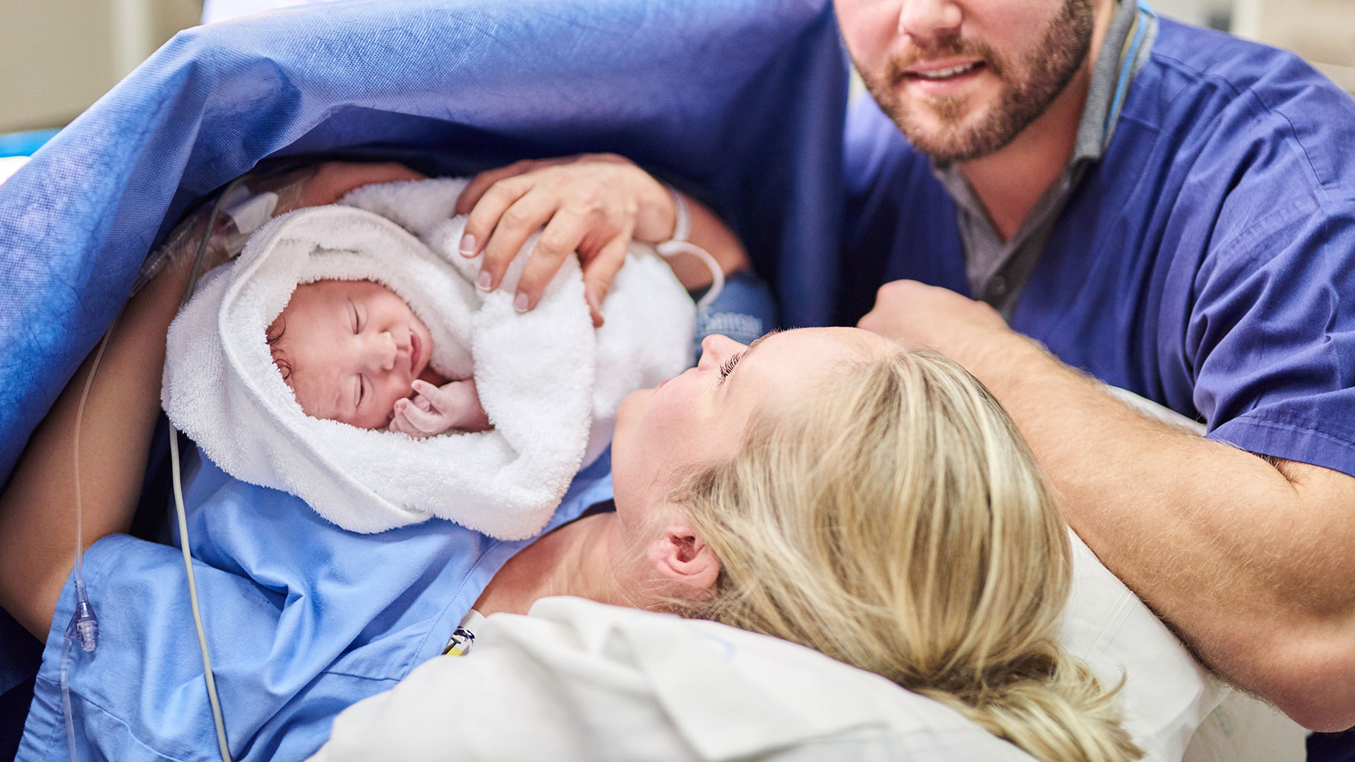 mulher deitada na cama em uma sala de cirurgia segura um bebê no colo. Ao lado dela, está um homem