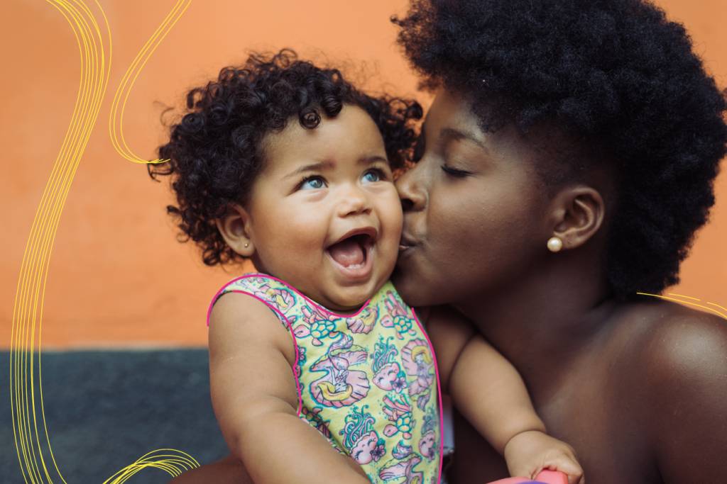 Mãe e filha, ambas de pele negra. A mãe está beijando a bochecha esquerda da menina, que tem por volta de um ano. A menina olha para cima e sorri com a boca aberta. O fundo da foto é laranja.