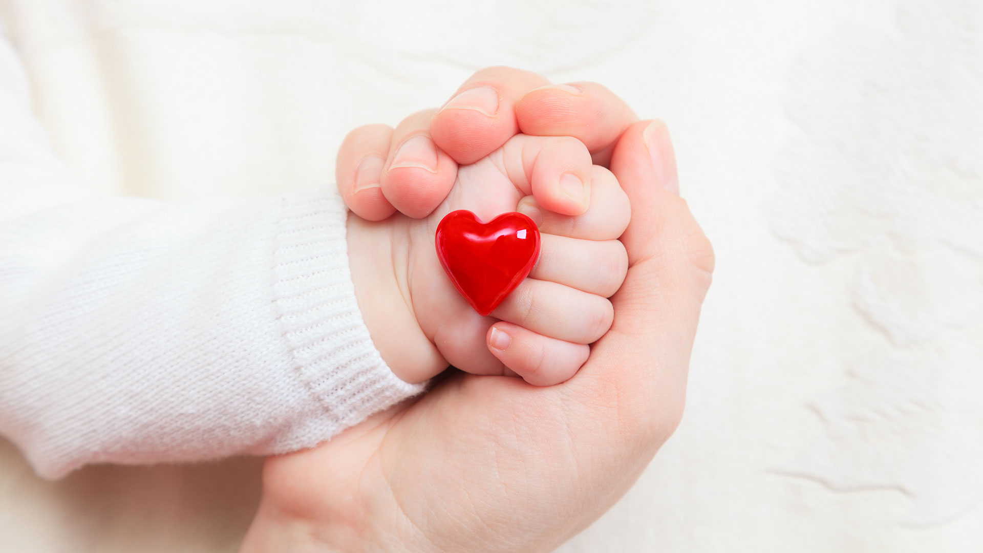 mão de um bebê envolta na mão de um adulto. O bebê tem um coração vermelho em frente os dedos