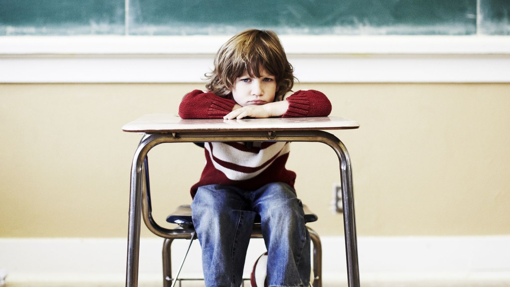 garoto sentado em uma carteira escolar, de costas para a lousa. Seu semblante é de chateação