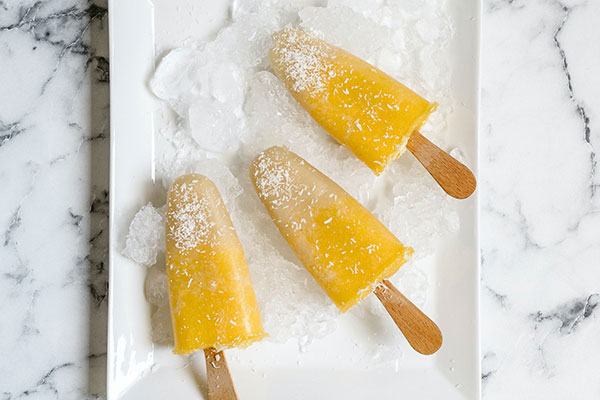 Three yellow popsicles with shredded coconut on top, on a white plate with ice