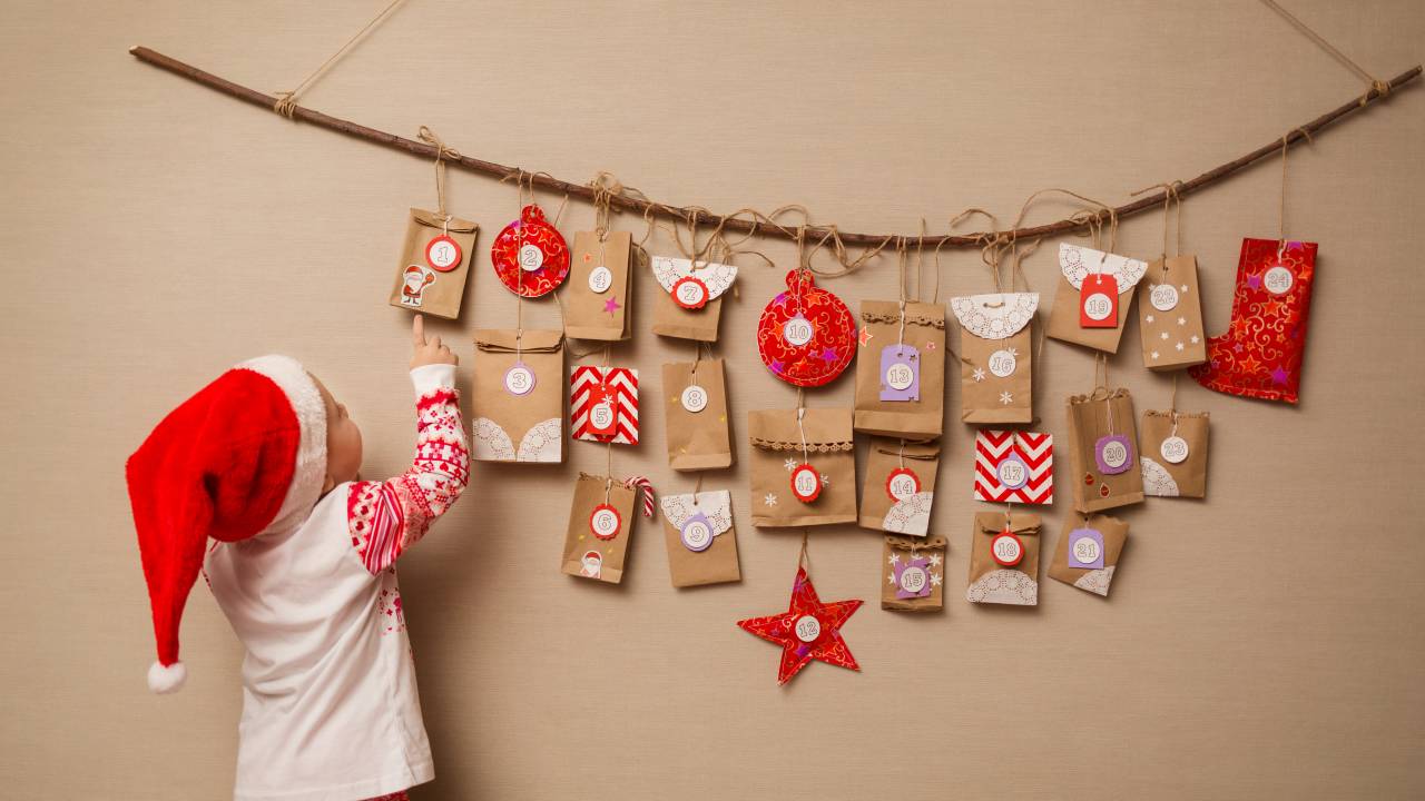 Criança de costas com roupa natalina vermelha e branca gorro de papai noel apontando para calendário do advento feito com saquinhos pendurados na parede