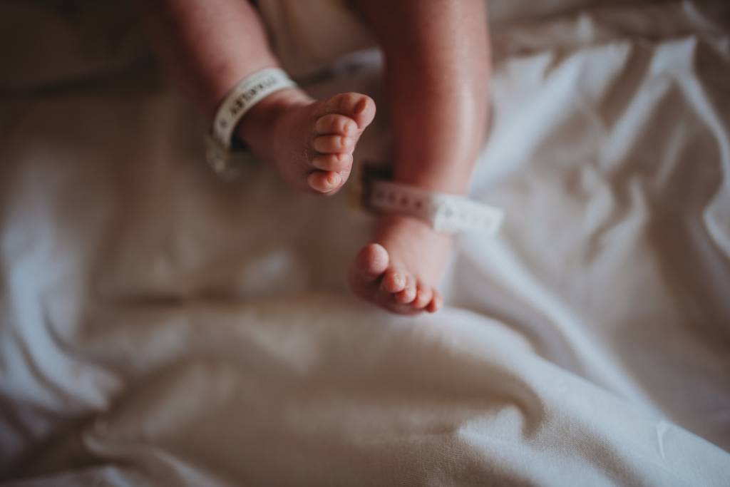 Panturrilhas e pés de um bebê recém-nascido, com identificação nas canelas. O lençol sobre o qual ele está é de cor branca. A pele do bebê é clara.