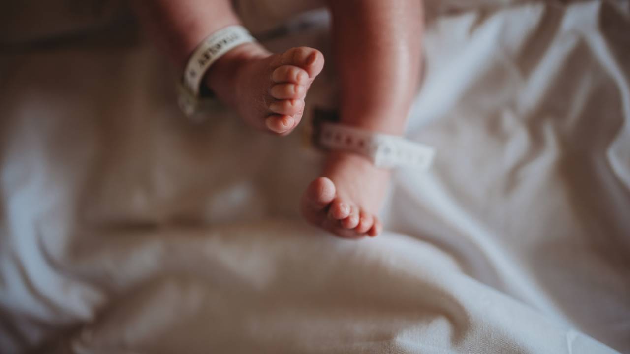 Panturrilhas e pés de um bebê recém-nascido, com identificação nas canelas. O lençol sobre o qual ele está é de cor branca. A pele do bebê é clara.