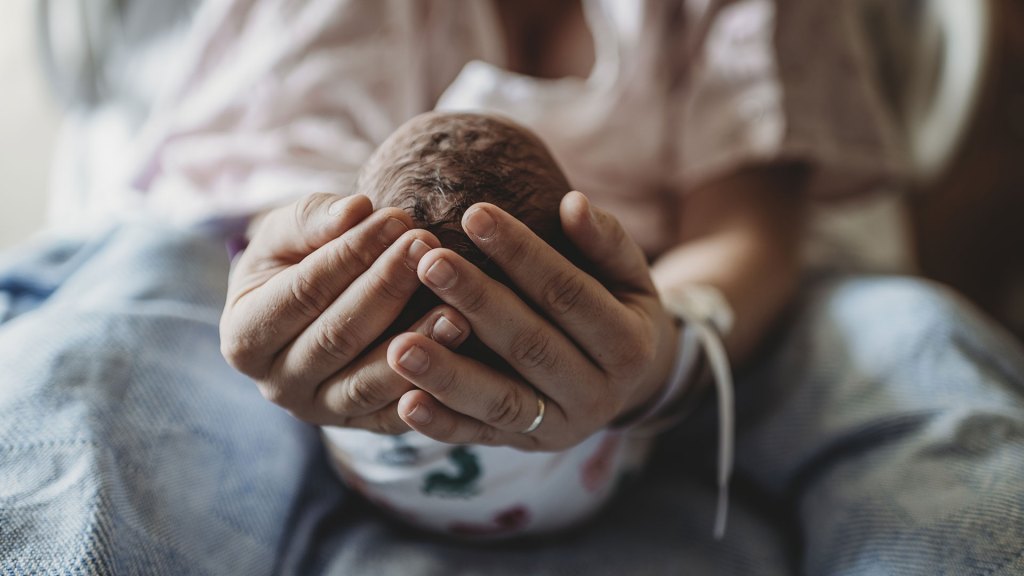 pessoa com as duas mãos encostadas segura a cabeça de um bebê em ambiente hospitalar