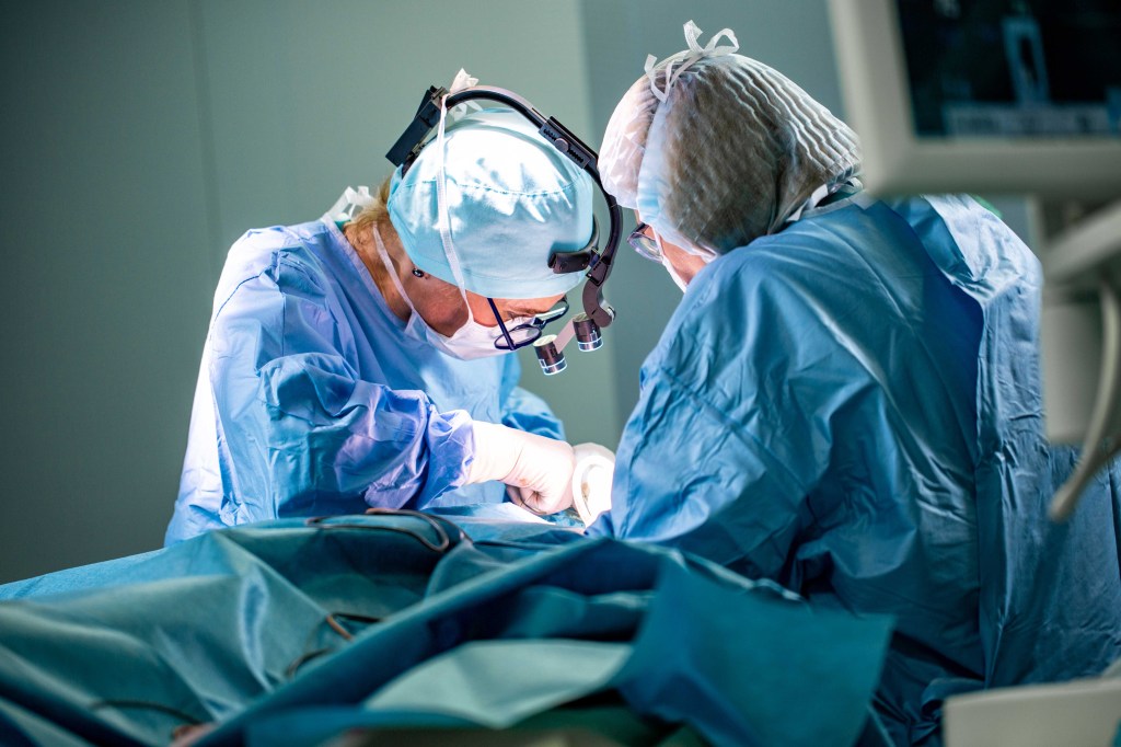 Na imagem, há cirurgiões com o uniforme azul, tucas e outros aparatos médicos realizando uma cirurgia.