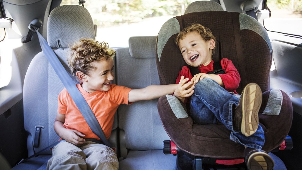 dois meninos sentados no banco de trás de um carro. Um deles está em uma cadeirinha. Eles sorriem olhando um para o outro