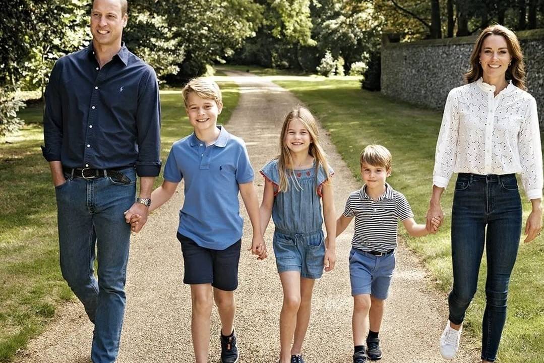 família real com william, george, charlotte, louis e kate. Estão todos de mãos dadas, nessa ordem, andando um lado do outro por um caminho em um parque.