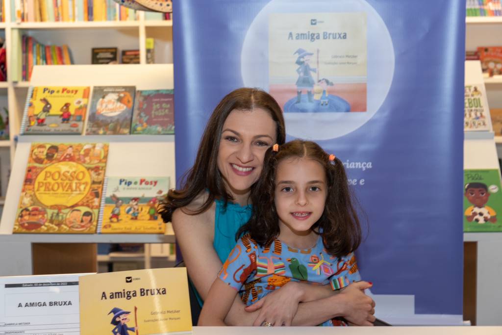 Gabriela Metzker, autora do livro, abraçando a filha no lançamento do livro. As duas estão sorrindo. Sobre a mesa, está o livro A Amiga Bruxa.