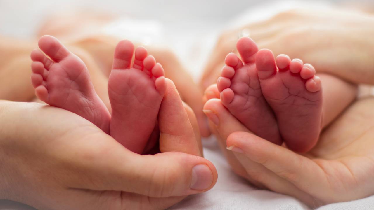 dois pares de pezinhos de bebês segurados por duas mãos adultas. Todos tem pele branca, os pezinnhos são rosados