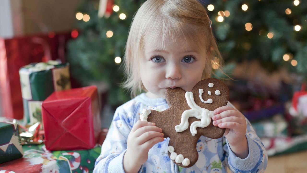 garota em frente a uma árvore de Natal cheia de presentes com um biscoito em forma de boneco na boca