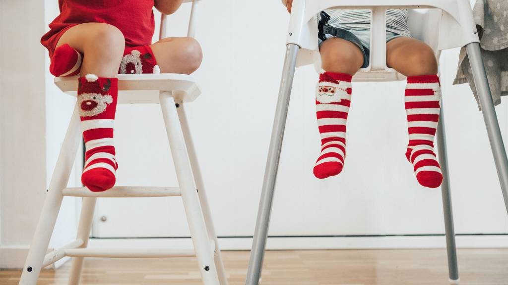 pernas de duas crianças sentadas em cadeirões de alimentação. Elas usam meias com temática de Natal