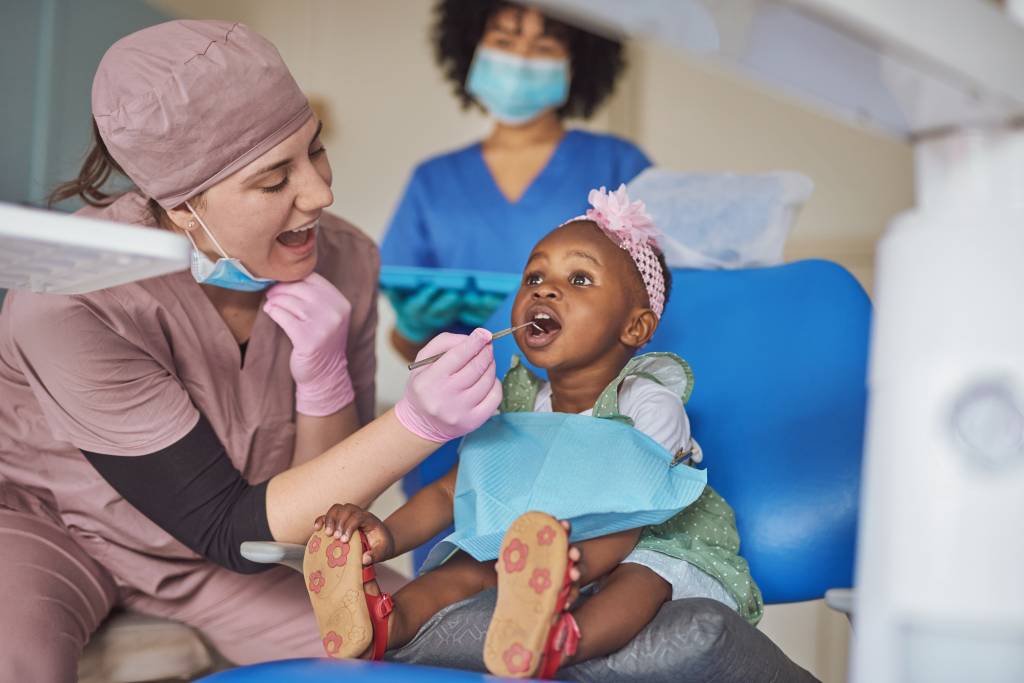 Criança em consulta ao dentista. Na foto, uma dentista com uniforme rosa escuro, luvas rosas, está sentada e atendendo uma criança negra. É uma menina com cerca de um ano, está com uma faixa rosa enfeitando a cabeça e com a boca aberta, onde a dentista coloca um instrumento. Ela não está chorando ou com medo.