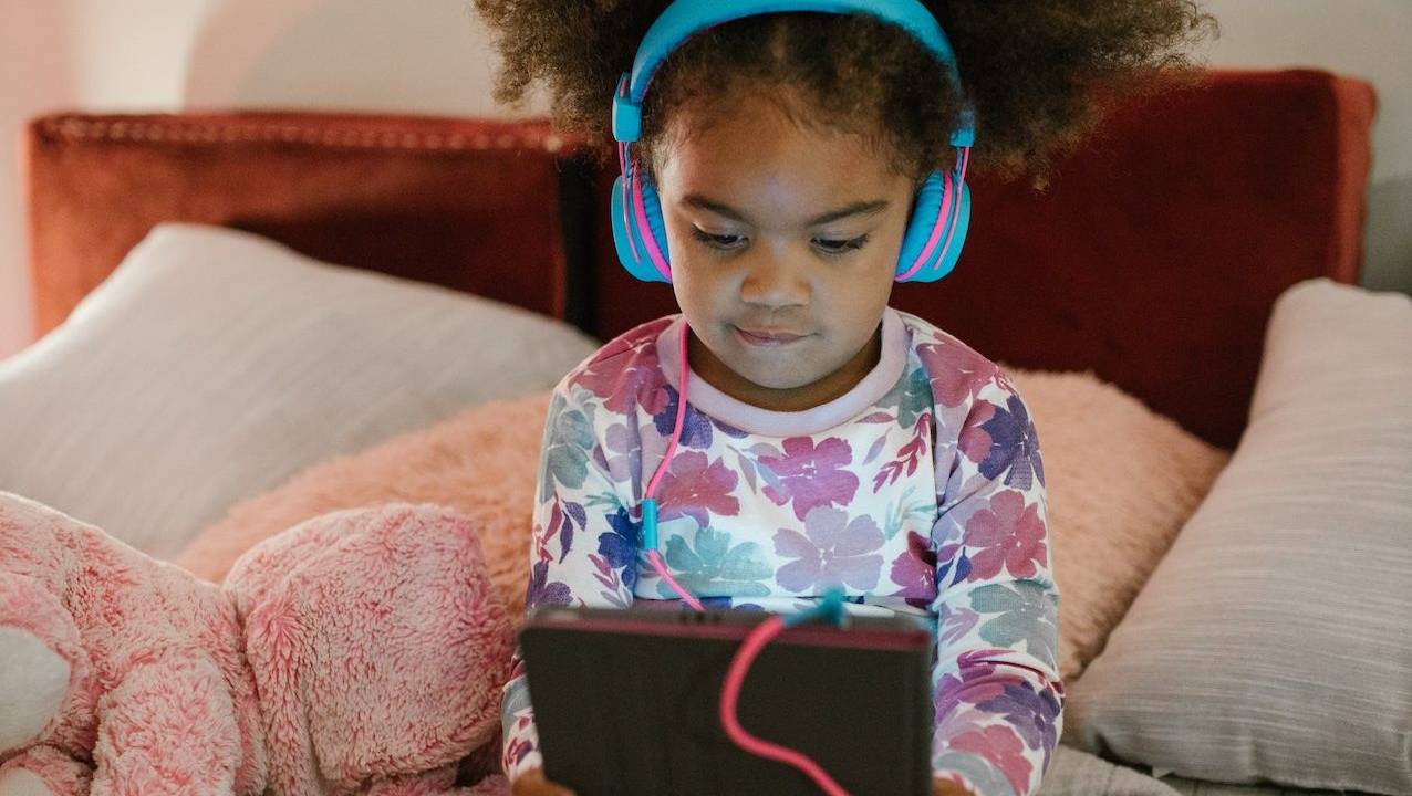 Criança jogando no tablet. É uma menina negra, sentada sobre a cama e vestindo pijama. Ela segura o tablet com as duas mãos e usa um grande fone de ouvido.