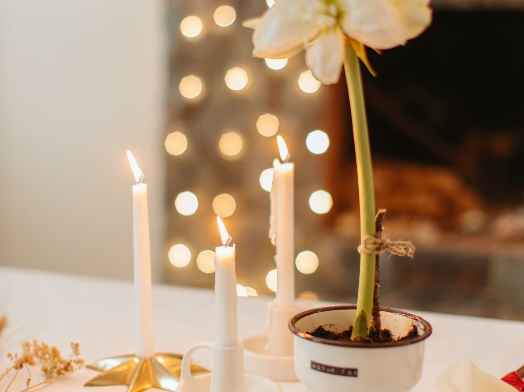 Três velas brancas sobre uma mesa branca, ao lado de um vaso de flores brancas. Ao fundo, é possível ver uma lareira com luzinhas de natal.