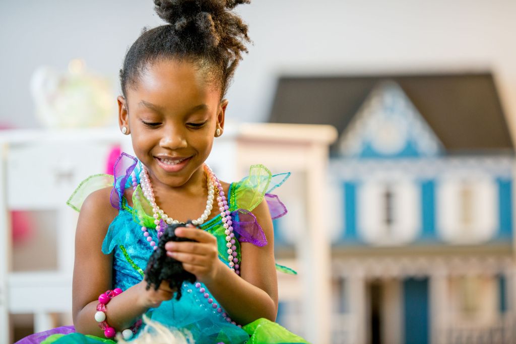 Menina negra, por volta de 5 anos, brincando com Barbie. Ela está de vestido azul com detalhes coloridos. Está com o cabelo preso e sorri olhando para a boneca.